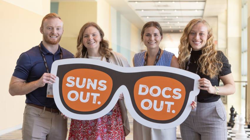 Four people standing in a row laughing hold a sign that looks like sunglasses that says Suns Out. Docs Out.