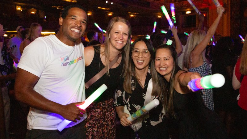 One man and three women smiling with glow sticks in a theater with others enjoying the party