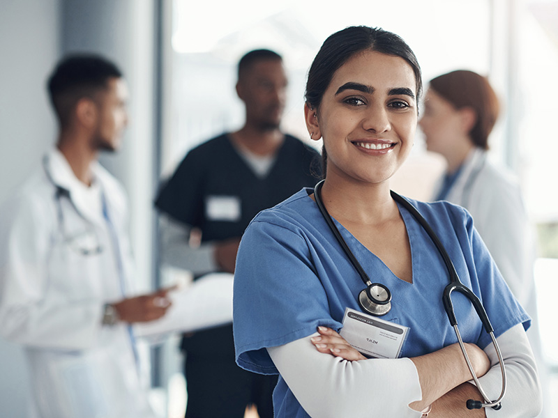 Female physician with stethescope around neck and arms crossed smiling with three colleagues in the background talking with each other.