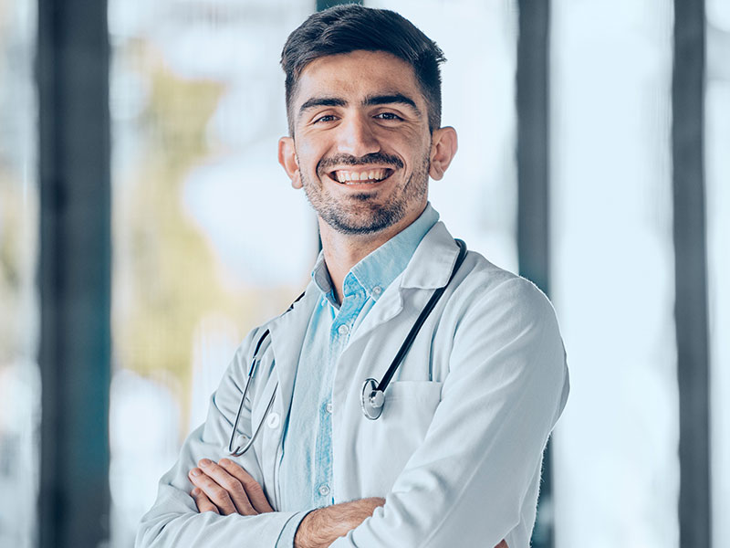 Young male doctor wearing white coat and stethoscope standing with arms cross smiling