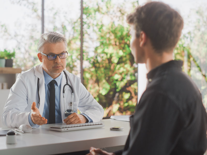 Physician sitting at desk speaking to young man sitting across the desk.